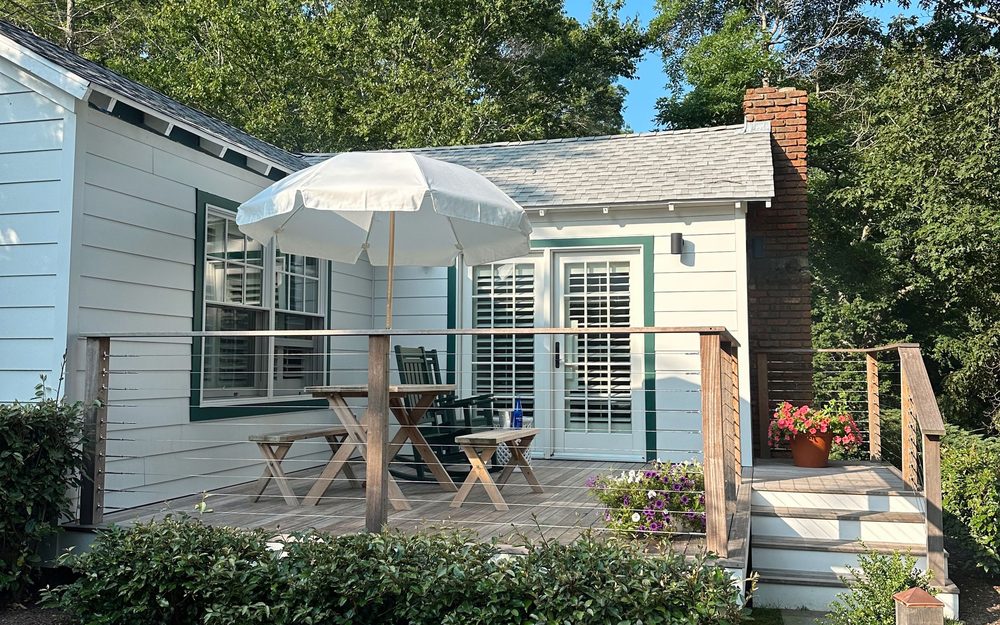 Small white cottage with French doors opening onto a wooden deck with a table, chairs and a patio umbrella