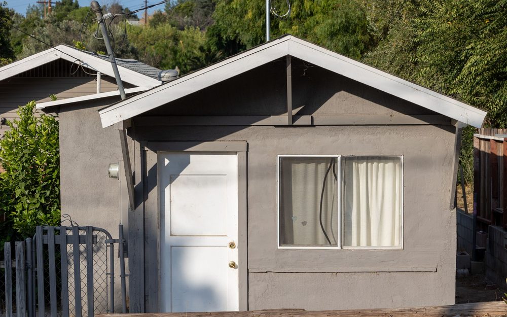 Small gray stucco outbuilding with a gable roof, a white door and a window, beside a wooden fence