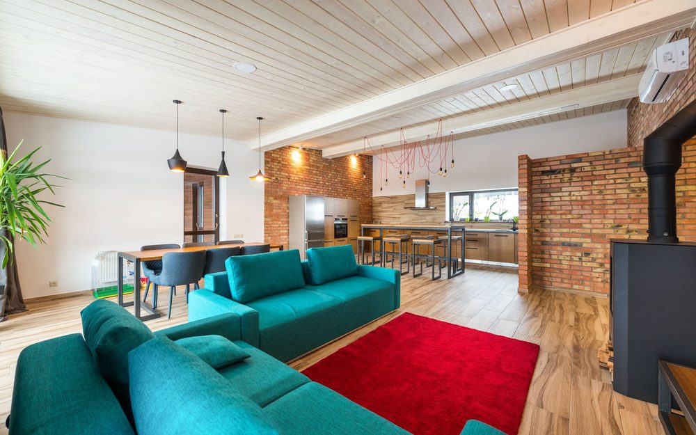 Open single-level living room with teal sofas, a red rug and a wood stove, opening onto a dining area and kitchen
