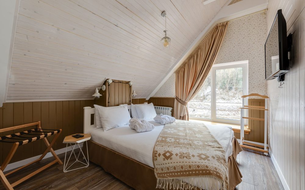 Bedroom with a sloped wood-paneled ceiling, a large window with curtains, and a bed with a patterned throw