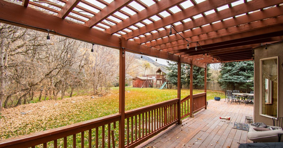 Long wooden deck with a railing and a pergola roof, looking out over a lawn and autumn trees