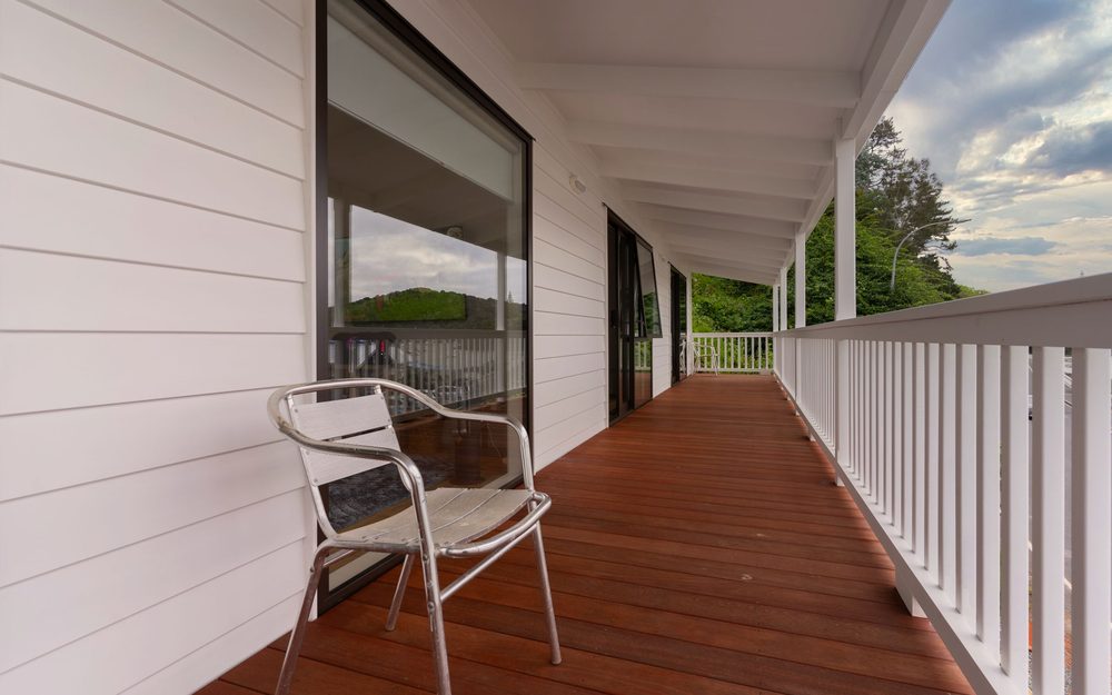 Long covered porch deck with reddish wood boards, a white railing and a chair, beside a white house