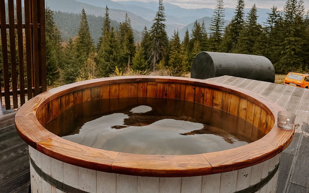 Round cedar hot tub on a wooden deck, with tall evergreen trees and hills behind
