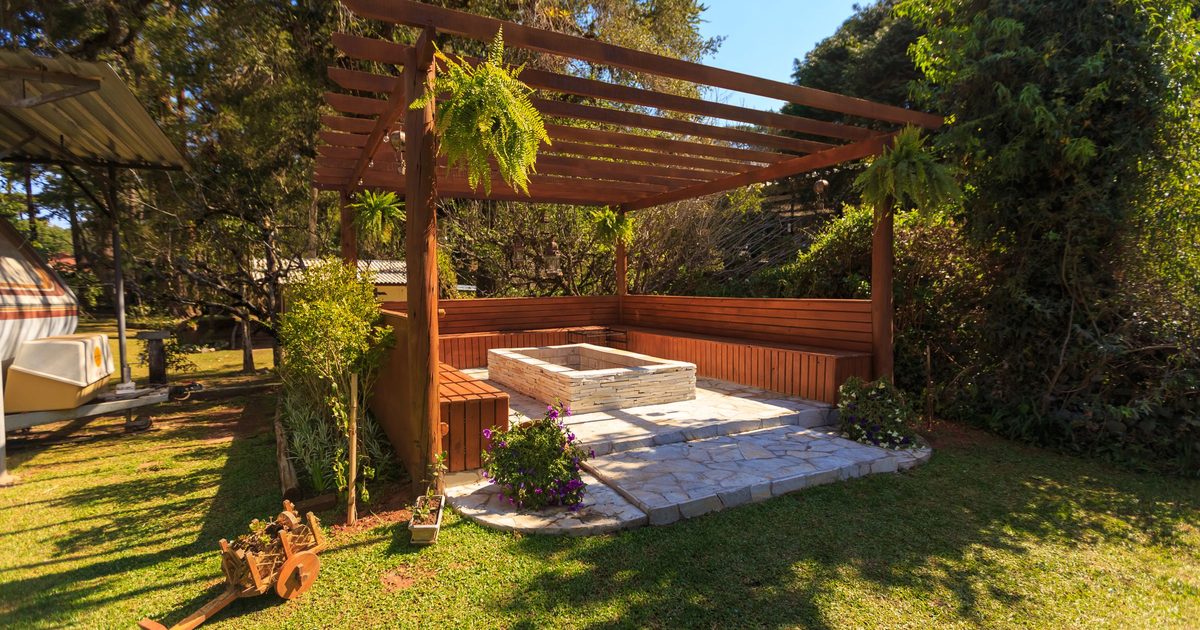 Wooden pergola over a stone patio with a built-in bench and a raised planter, in a green backyard with trees