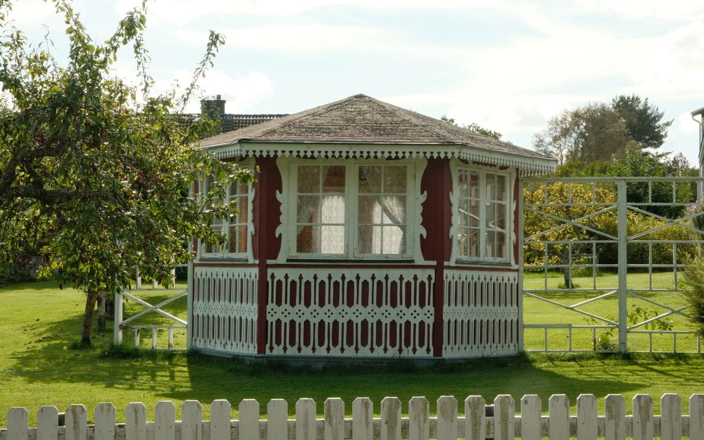 White and red octagonal gazebo with a shingled roof, standing in a grassy yard with fruit trees