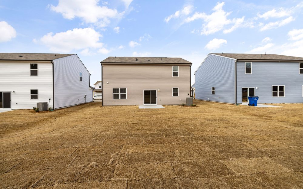 Open backyard of bare ground between the backs of three houses, under a blue sky with clouds