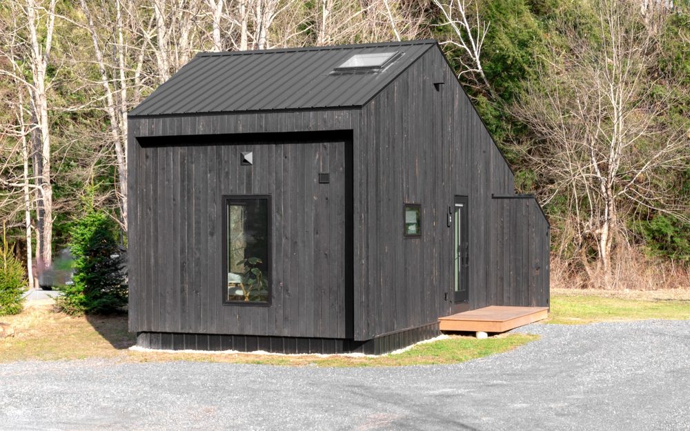 Small black wood-clad cabin with a metal roof and a skylight, on gravel among bare trees