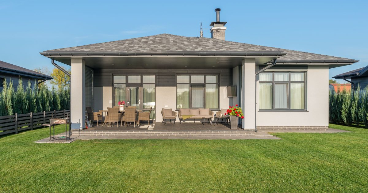 Single-story house with a covered patio built into the main roofline, outdoor dining and lounge furniture underneath, and gutters and downspouts at each corner