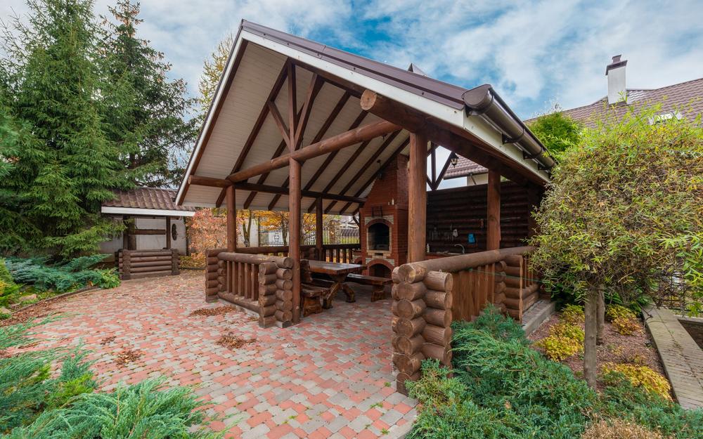 Gable roof patio cover with exposed timber beams over a brick patio, with evergreens behind