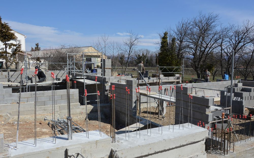 Concrete block foundation walls with steel bars being built on a cleared site, with trees and houses behind