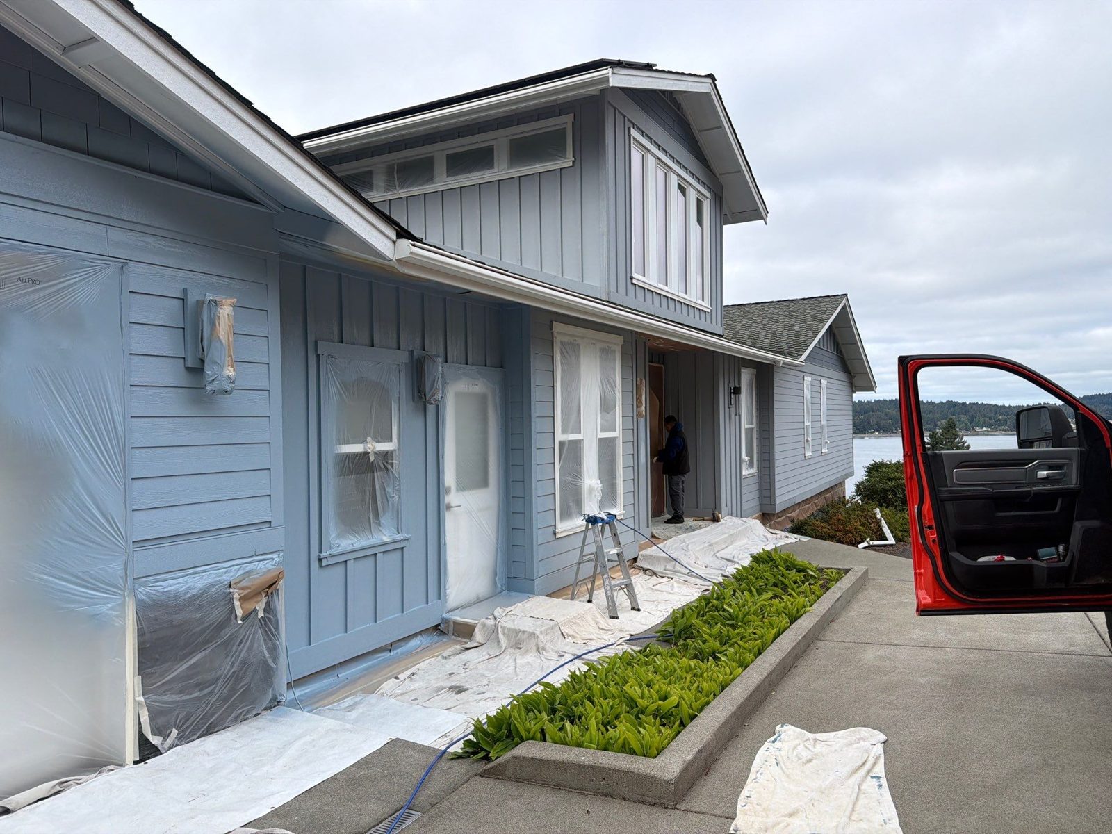 Crew repainting the board-and-batten siding of a waterfront house, windows masked and drop cloths down